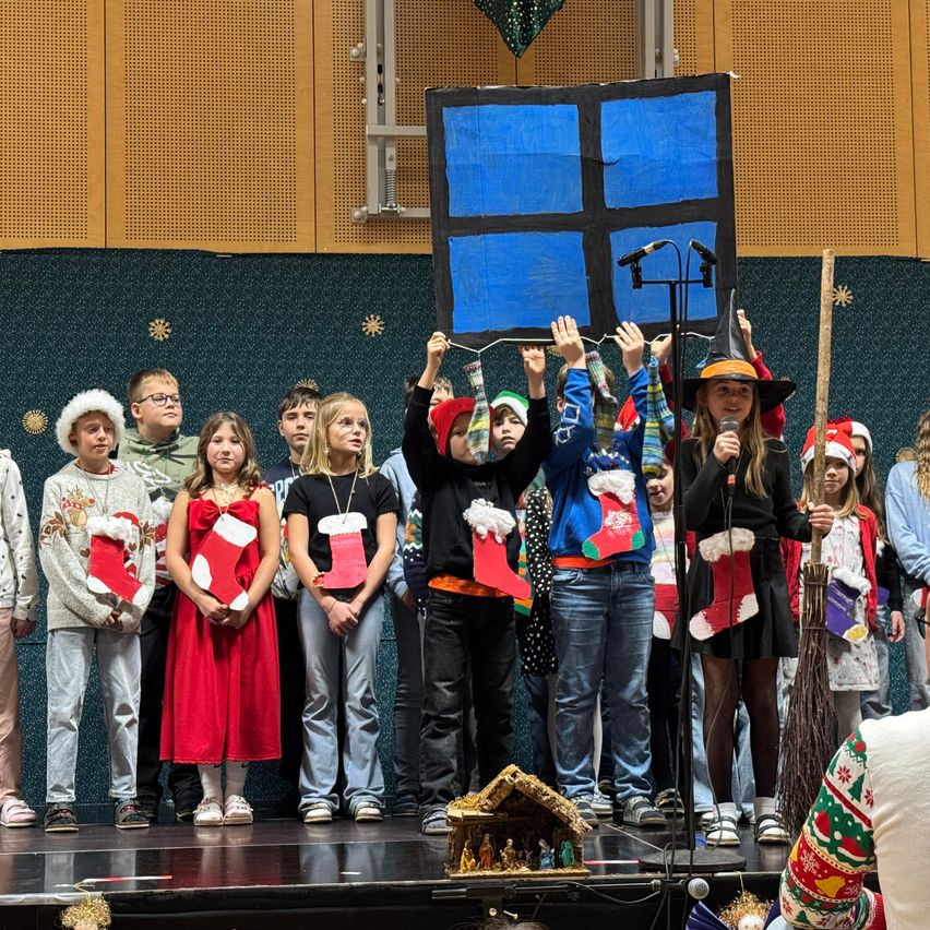 Eine Gruppe von Kindern in festlicher Kleidung, einige mit roten Mützen, steht auf der Bühne und hält Weihnachtssocken. Ein Mädchen mit Hut hält ein Mikrofon und ein blaues Quadrat ist über ihnen positioniert.