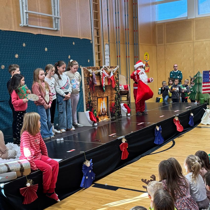 Eine Gruppe von Kindern sieht eine Aufführung von Santa Claus auf einer Bühne, die mit weihnachtlichen Themen und amerikanischen Flaggen dekoriert ist. Der Weihnachtsmann steht, hält einen Sack und scheint eine Show aufzuführen.