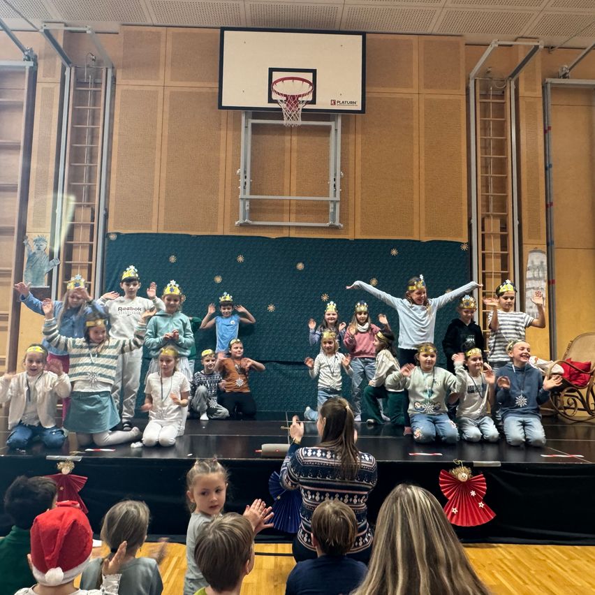 Eine Gruppe von Kindern mit Kronen auf dem Kopf tritt auf einer Bühne mit grünem Hintergrund auf. Eine Frau steht vor ihnen. Zuschauer sehen von den Tribünen zu, einige tragen festliche Mützen.
