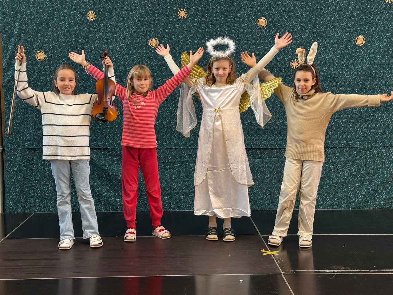 Fünf Mädchen stehen auf einer Bühne und lächeln. Drei von ihnen tragen Engelsflügel und Hasenohren, während zwei lässige Kleidung tragen. Eines hält eine Geige.