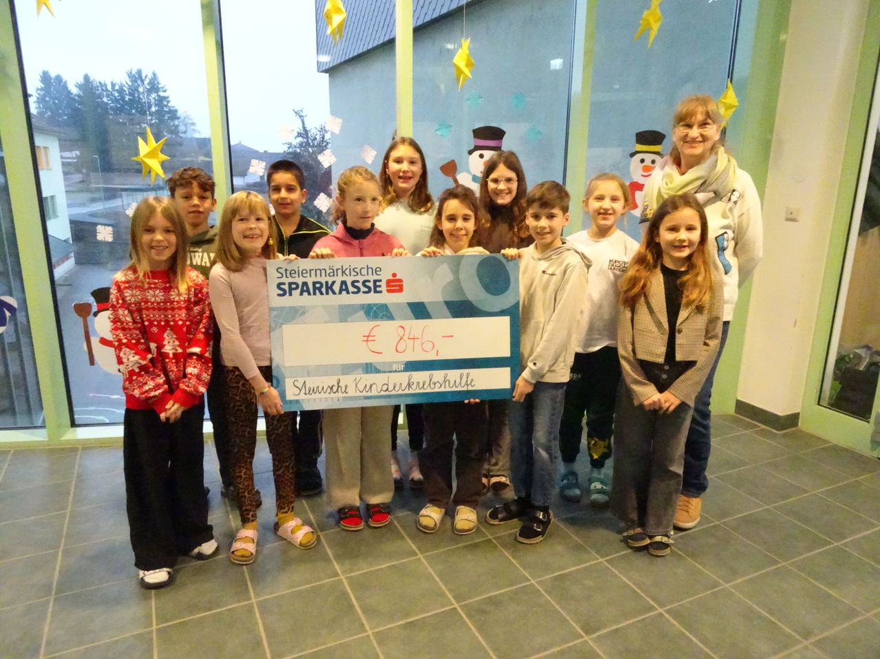 Eine Gruppe von Kindern und Erwachsenen posiert mit einem großen Scheck von der Steiermärkische Sparkasse für Kinderkrebshilfe. Sie stehen in einem Raum mit festlicher Weihnachtsdekoration.