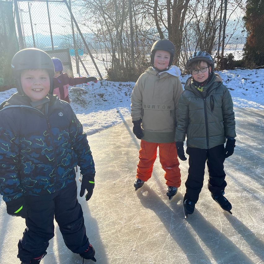 Three children stand on a snowy ice rink, all wearing helmets and winter clothing. Two of them are wearing sneakers while the third has on orange pants. They are smiling, and a fence with trees is behind them.