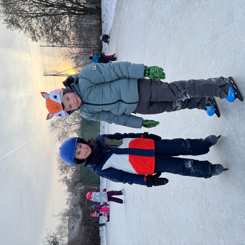 Two children in winter clothes skate on an ice rink with a clear sky and distant trees in the background.