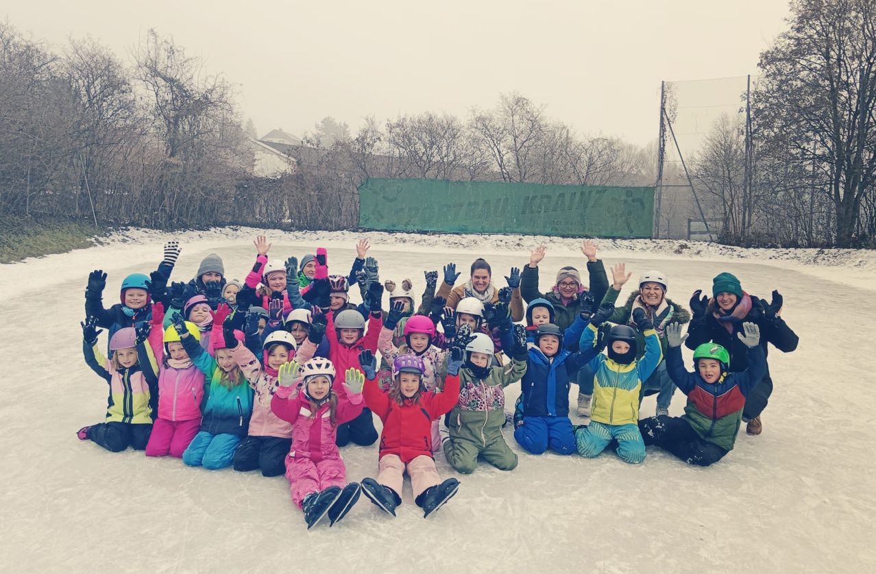 A group of children and adults in winter clothing pose for a photo on an ice rink. They are all smiling and some are raising their hands.