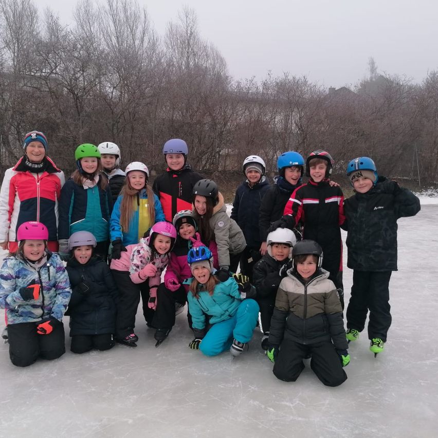A group of children in winter gear, helmets, and skates pose for a photo on an ice rink, with trees and a foggy sky in the background.