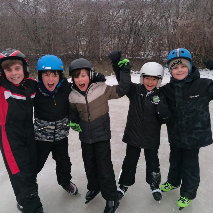 Five boys wearing winter gear and helmets are standing on ice. They are smiling and posing for a photo. The boys are wearing gloves and sneakers. They are all wearing different colored helmets.