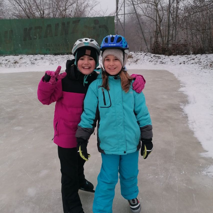 Two girls are standing on an ice rink, wearing winter clothes and helmets. The girl on the left is smiling and making a peace sign. Behind them is a snow-covered area.