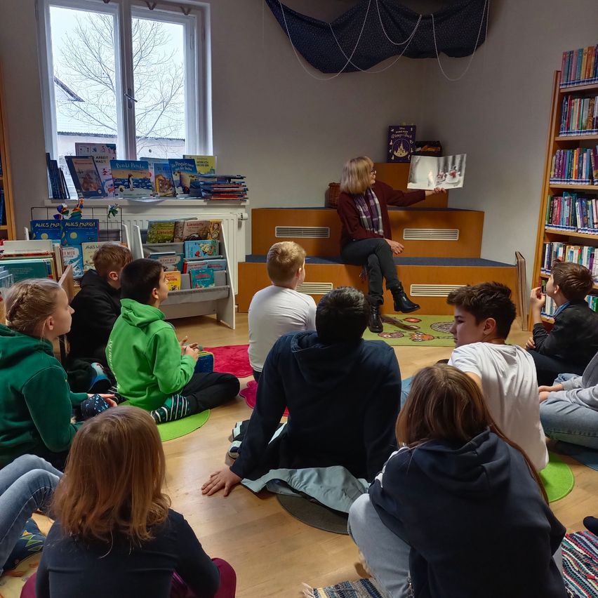 Eine Frau sitzt auf hölzernen Stufen in einer Bibliothek, hält ein Buch in der Hand und liest einer Gruppe von Kindern vor, die im Kreis auf dem Boden sitzen. Der Raum hat Bücherregale und ein Fenster.