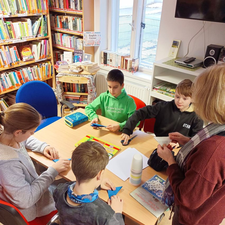 Eine Gruppe von Kindern arbeitet in einer Bibliothek an einem Projekt. Eine Frau in einem roten Pullover hilft ihnen.