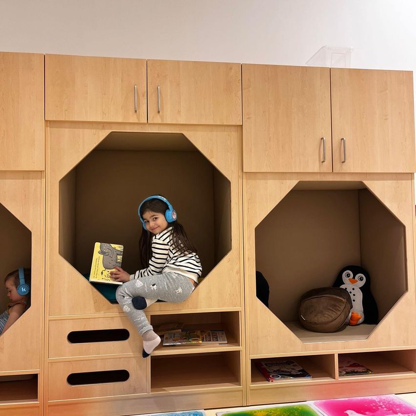 Zwei junge Mädchen sitzen in einem sechseckigen, hölzernen Versteck in einem Spielzimmer. Ein Mädchen trägt Kopfhörer und liest ein Buch, während das andere Kopfhörer trägt und zu schlafen scheint.