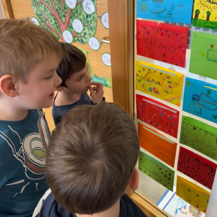 Drei junge Jungen betrachten bunte Karten mit Namen und Zeichnungen an einer Wand. Die Karten haben verschiedene Farben und Designs. Die Wand ist mit einem Baum und Buchstaben des Alphabets dekoriert.