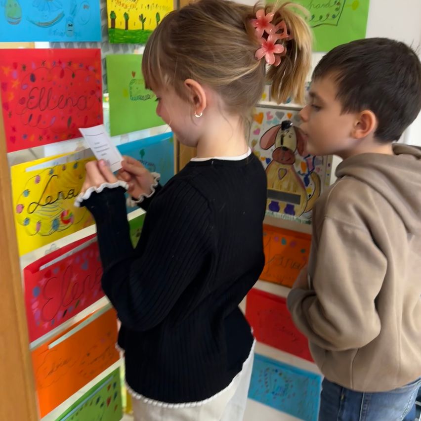 Ein Mädchen liest ein Papier, während es neben einem Jungen steht, der ihr ins Ohr bläst. Hinter ihnen hängen farbenfrohe Zeichnungen an der Wand.