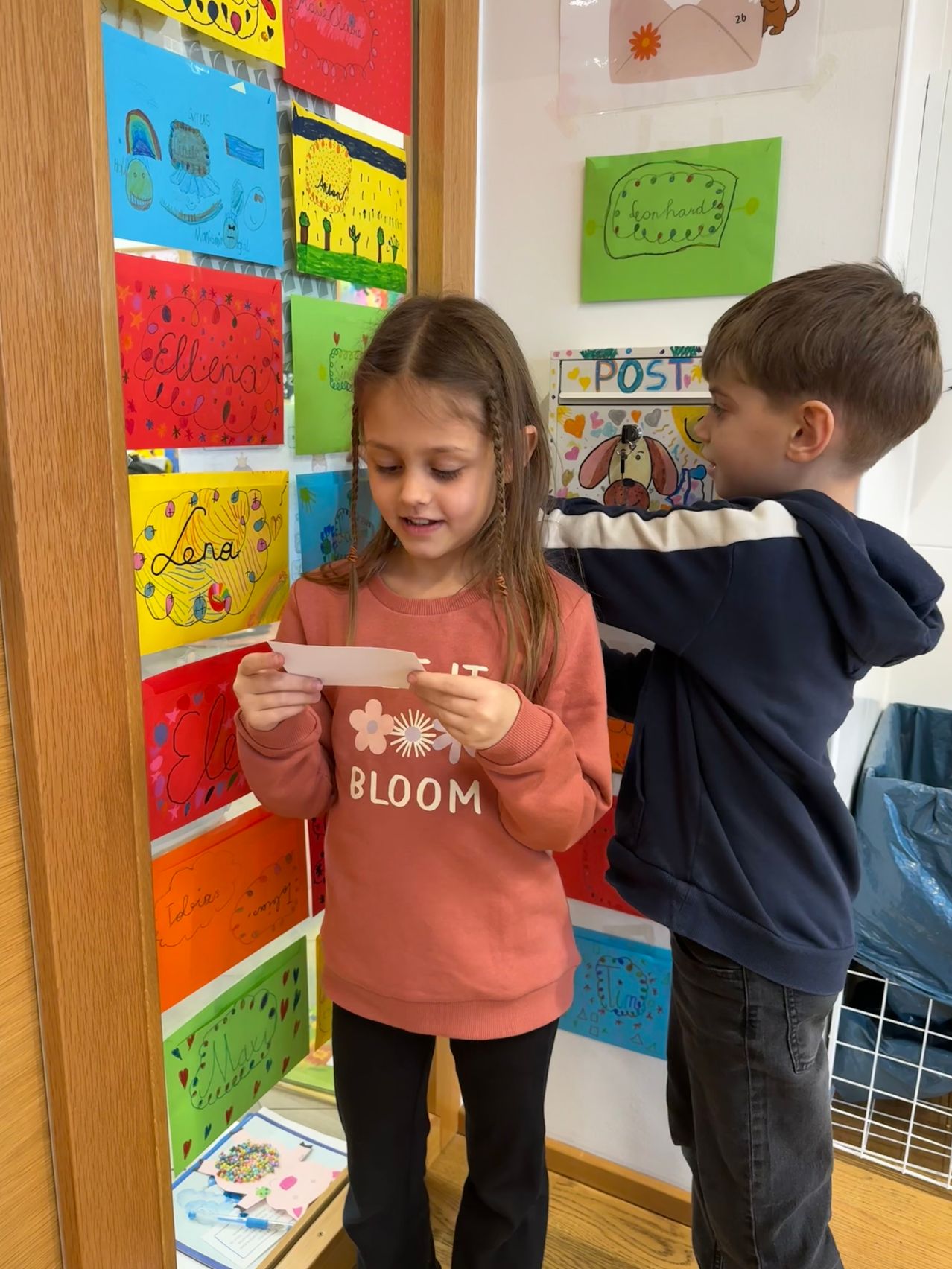 Zwei Kinder stehen vor einer Pinnwand mit bunten Zeichnungen. Ein Mädchen hält ein Papier und ein Junge schaut sie an.