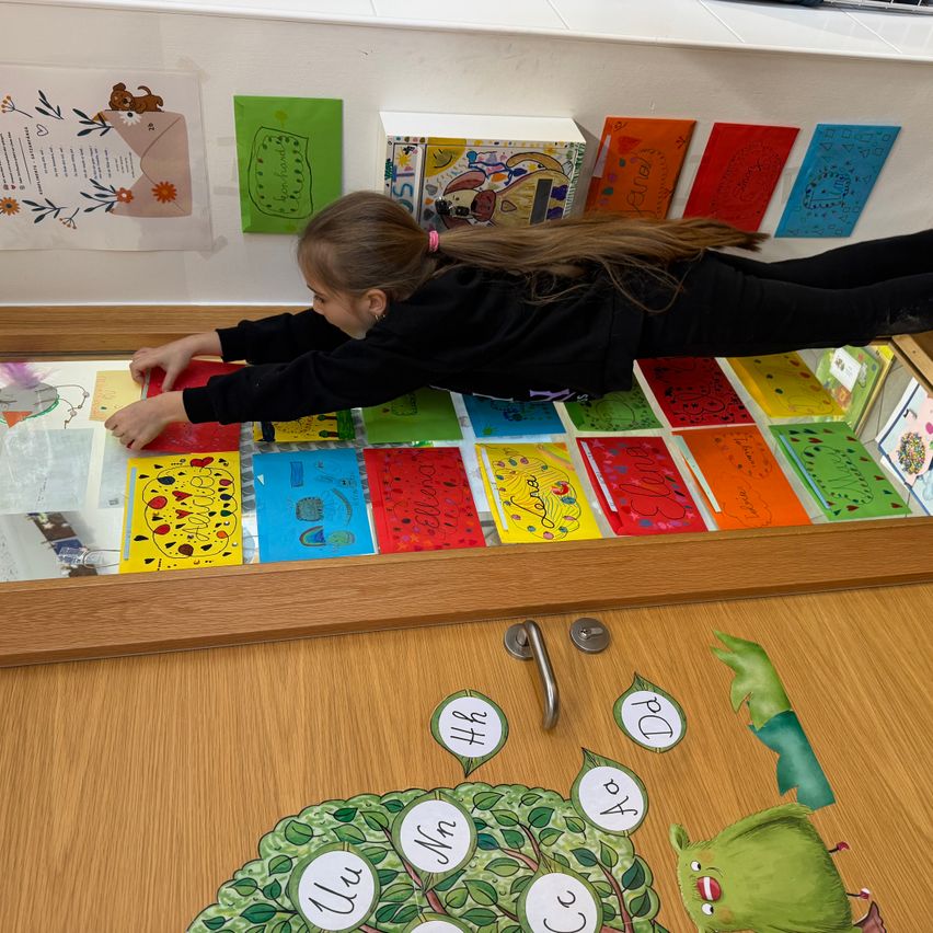 Ein junges Mädchen liegt auf dem Boden vor einer Glasvitrine mit bunten Karten. Die Karten haben Zeichnungen und Text darauf. Die Wand hinter ihr hat verschiedene farbige Poster und eine Box.