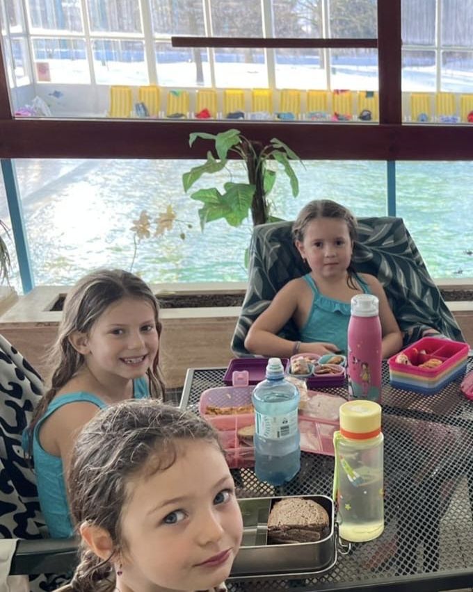 Drei junge Mädchen sitzen lächelnd für die Kamera an einem Tisch. Hinter ihnen befindet sich ein Schwimmbecken mit Blick auf einen Wasserpark. Mittagessen und Flaschen stehen auf dem Tisch.