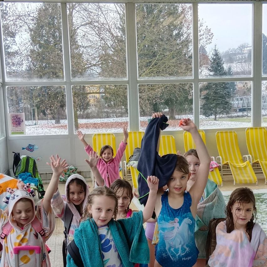 Eine Gruppe junger Mädchen steht in einem Schwimmbereich, einige heben die Hände. Sie tragen Handtücher und einige sind in Badekleidung. Hinter ihnen stehen leere gelbe Stühle und draußen ist die Aussicht auf Bäume.