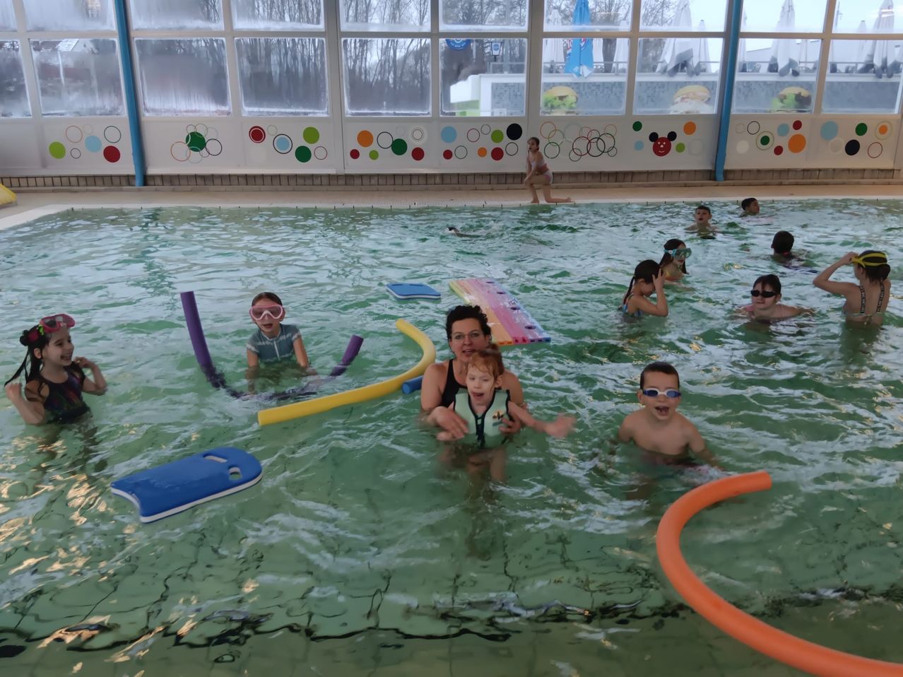 Eine Gruppe von Kindern und Erwachsenen schwimmt im Pool, einige tragen Schwimmbrillen und Schwimmwesten. Bunte Kreise schmücken die Wände.
