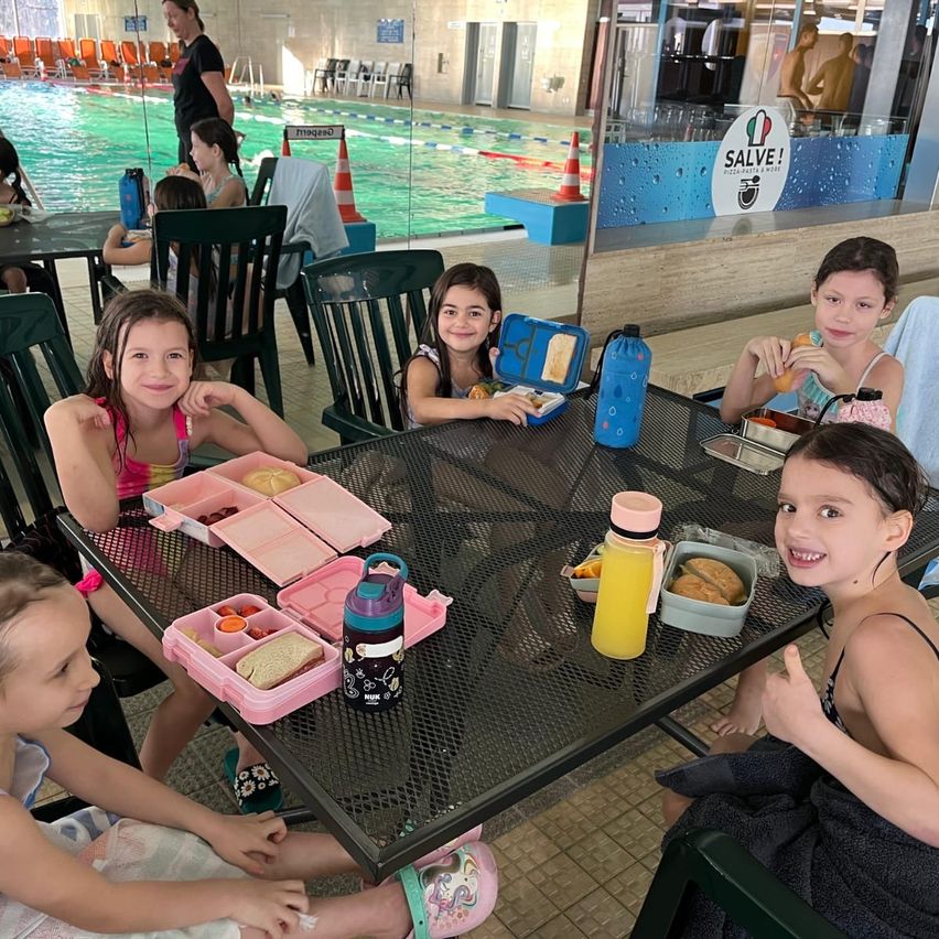 Fünf junge Mädchen sitzen an einem Tisch im Schwimmbadbereich und essen ihr Mittagessen. Jedes Mädchen hat einen Tablett mit Essen und Getränken. Hinter ihnen befindet sich ein Pool, eine Bank und ein Schild. Eine Frau steht in der Nähe des Pools.
