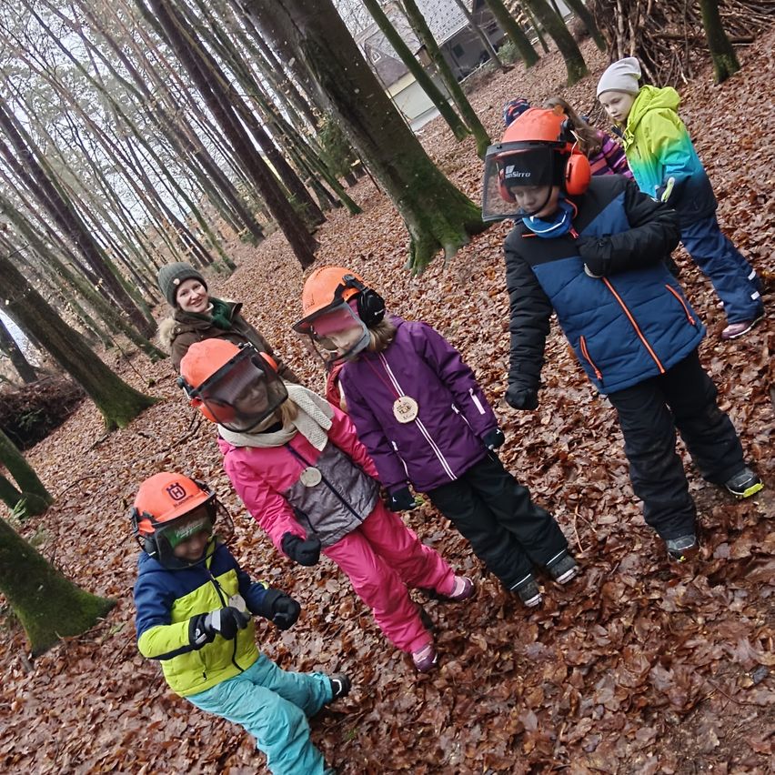 Eine Gruppe von Kindern im Wald, in Winterkleidung und orangefarbenen Helmen, steht in einer Reihe. Sie scheinen an einer Winteraktivität beteiligt zu sein, möglicherweise Wandern oder eine Schnitzeljagd.