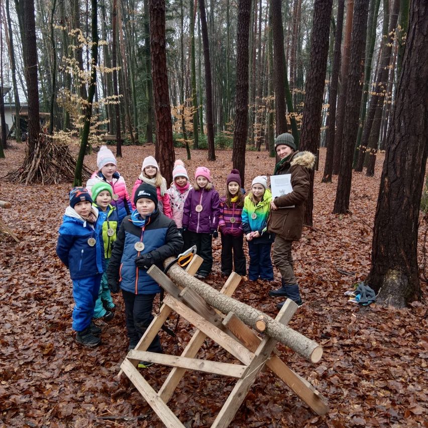 Eine Gruppe von Kindern, alle in Winterkleidung, steht in einem Wald. Sie posieren für ein Foto mit einem Erwachsenen, der ein Papier in der Hand hält.