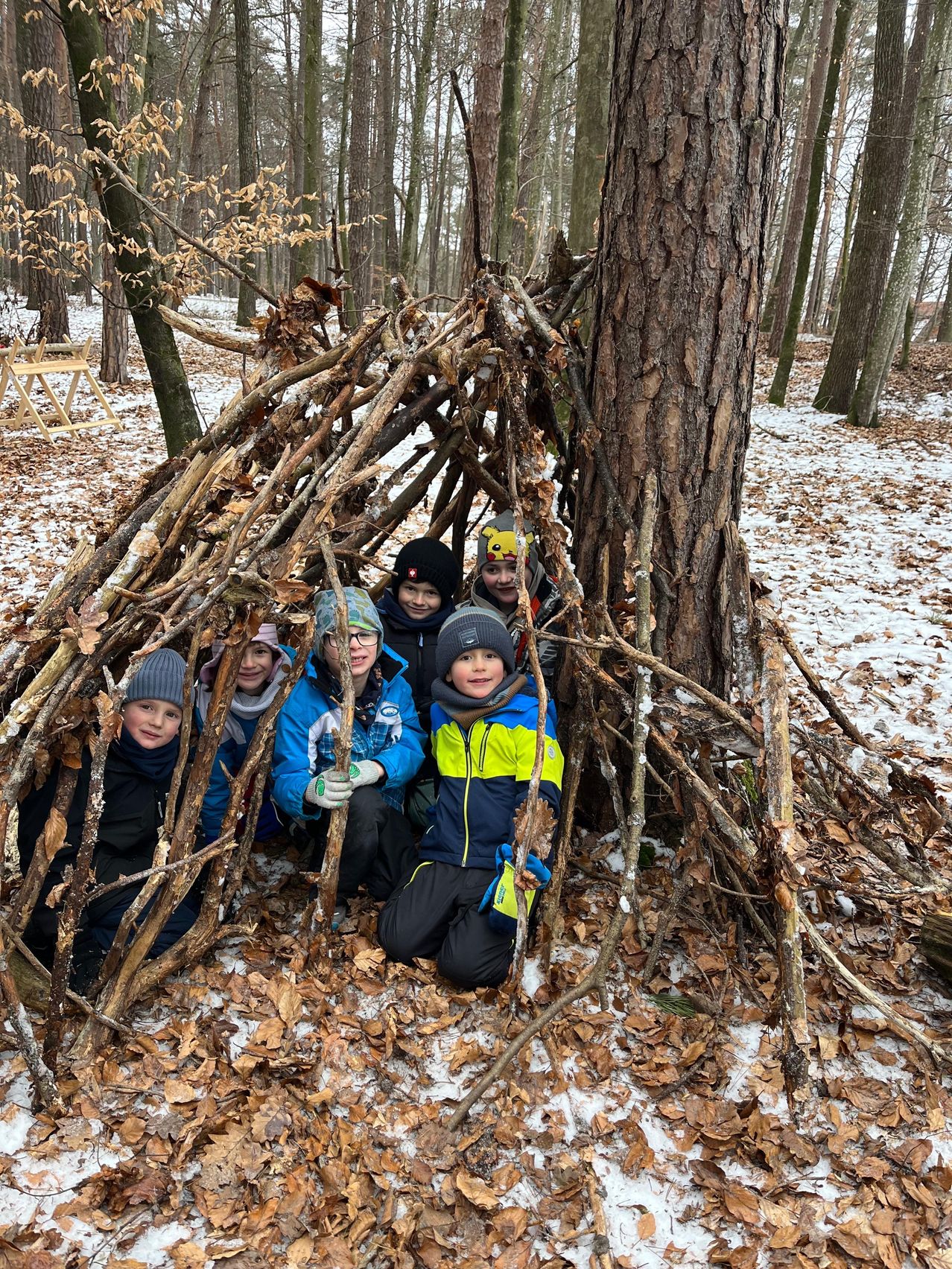 Eine Gruppe von Kindern posiert in einem von Ästen gebauten Unterschlupf in einem verschneiten Wald. Der Unterschlupf wird von einem hohen Baumstamm gestützt.