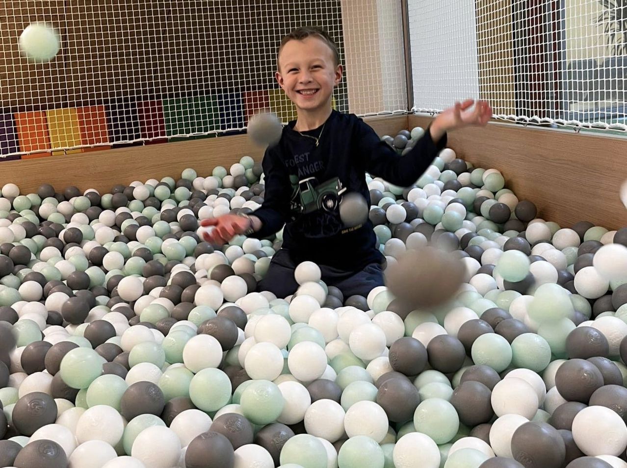 Ein junger Junge sitzt in einem Ballpool voller weißer und grauer Bälle und lächelt. Er trägt ein schwarzes Shirt.
