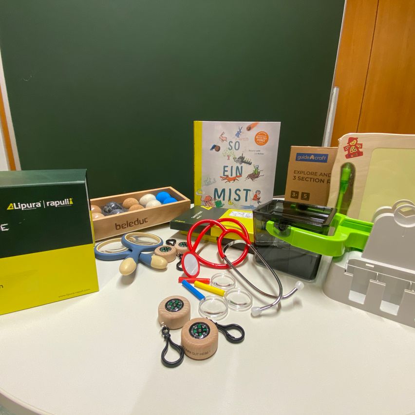 Ein Tisch mit Lernspielzeug und Büchern. Ein Kompass, ein Stethoskop und andere Werkzeuge sind darauf verstreut. Ein Buch mit dem Titel 'So ein Mist' ist vorhanden.
