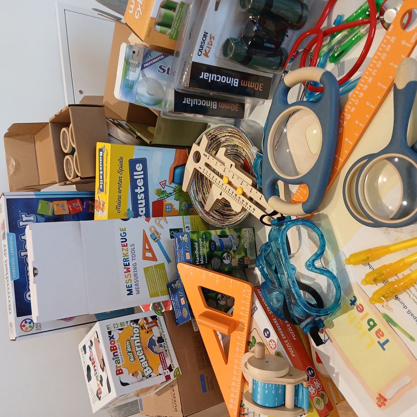 Eine Sammlung verschiedener pädagogischer und handwerklicher Werkzeuge, darunter Messwerkzeuge, Scheren, Lineale und Schutzbrillen, auf einem Tisch angeordnet.