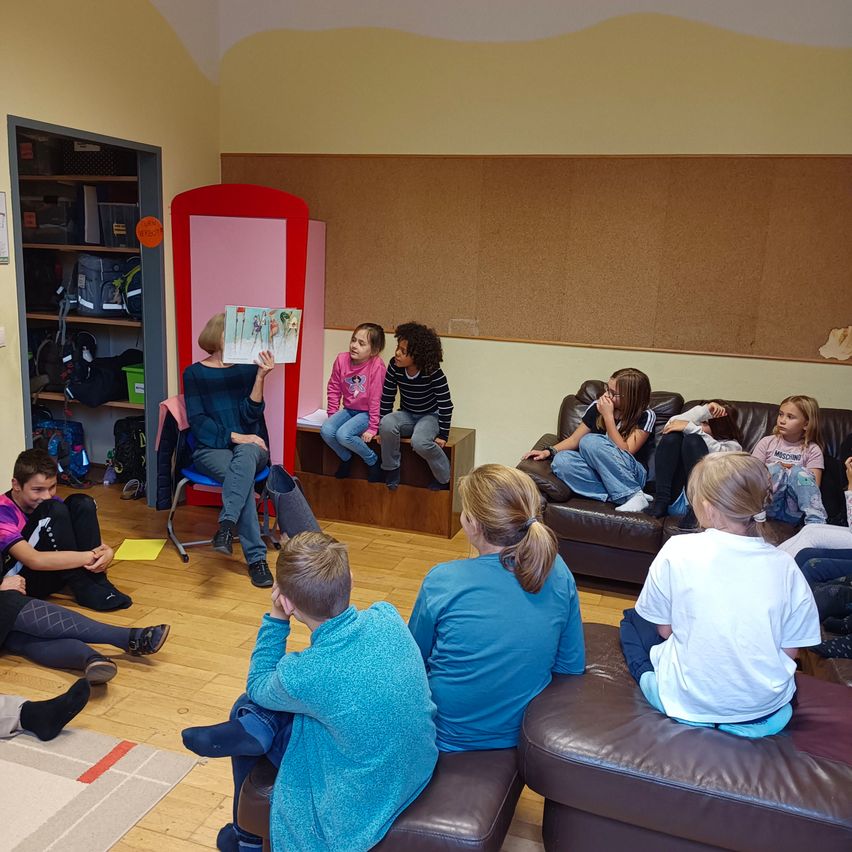 Eine Frau liest ein Buch vor einer Gruppe von Kindern, die auf einer Couch und dem Boden in einem Raum mit Holzboden und einer braunen Wand sitzen.