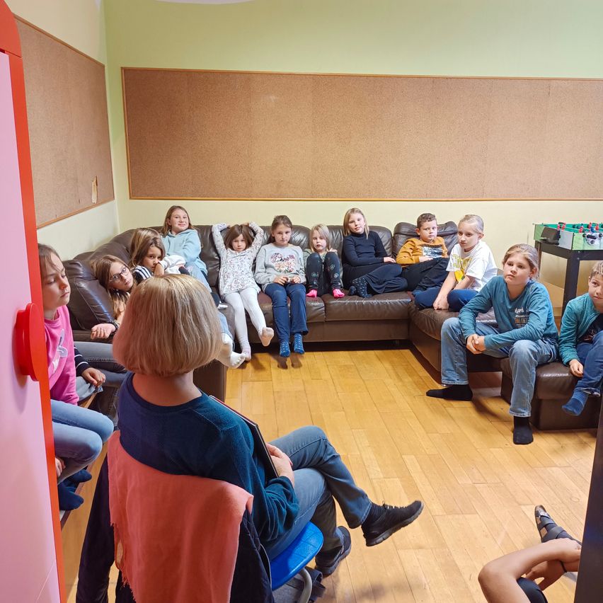 Kinder sitzen auf einer Couch im Klassenzimmer, einige stehen. Ein Lehrer sitzt vorne mit einem Buch. Dahinter hängt eine Korktafel an der Wand.