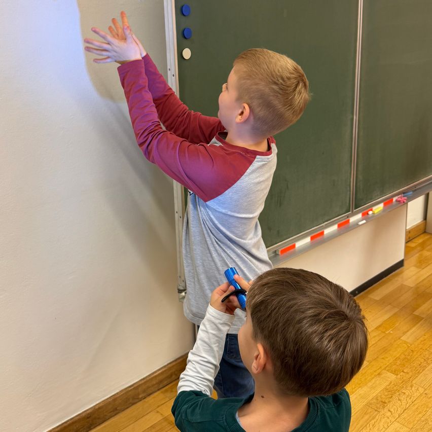 Two young boys are standing in a classroom, one with his hands raised and the other holding a blue object. They are in front of a green board with various colored buttons.