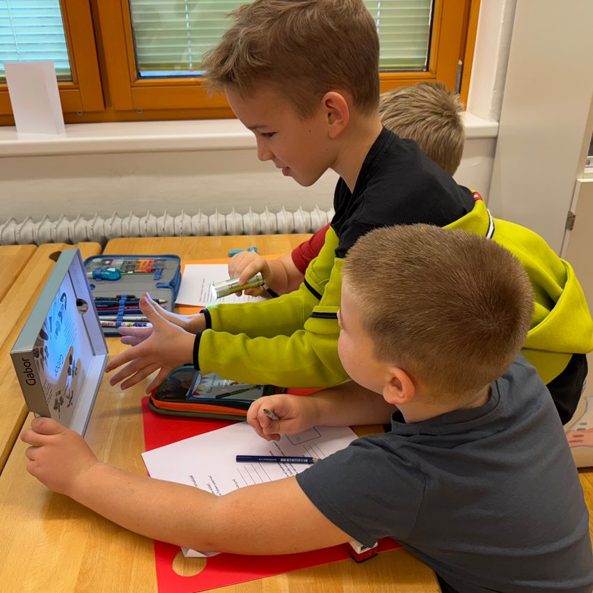 Three boys are seated at a table in a classroom. One boy is pointing at a screen, while another writes on a piece of paper. A box of crayons is on the table.