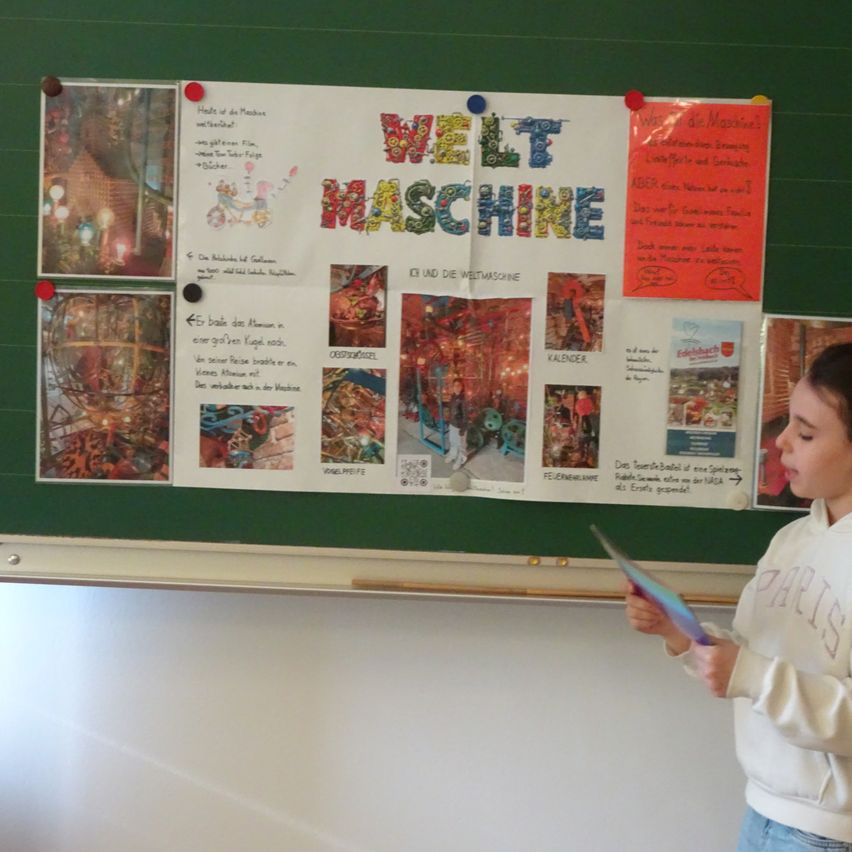 Ein Schüler liest ein Buch im Klassenzimmer und steht vor einer grünen Pinnwand mit verschiedenen Postern und Bildern. Ein Poster trägt den Titel 'Welt Maschine' und zeigt farbenfrohe Illustrationen.
