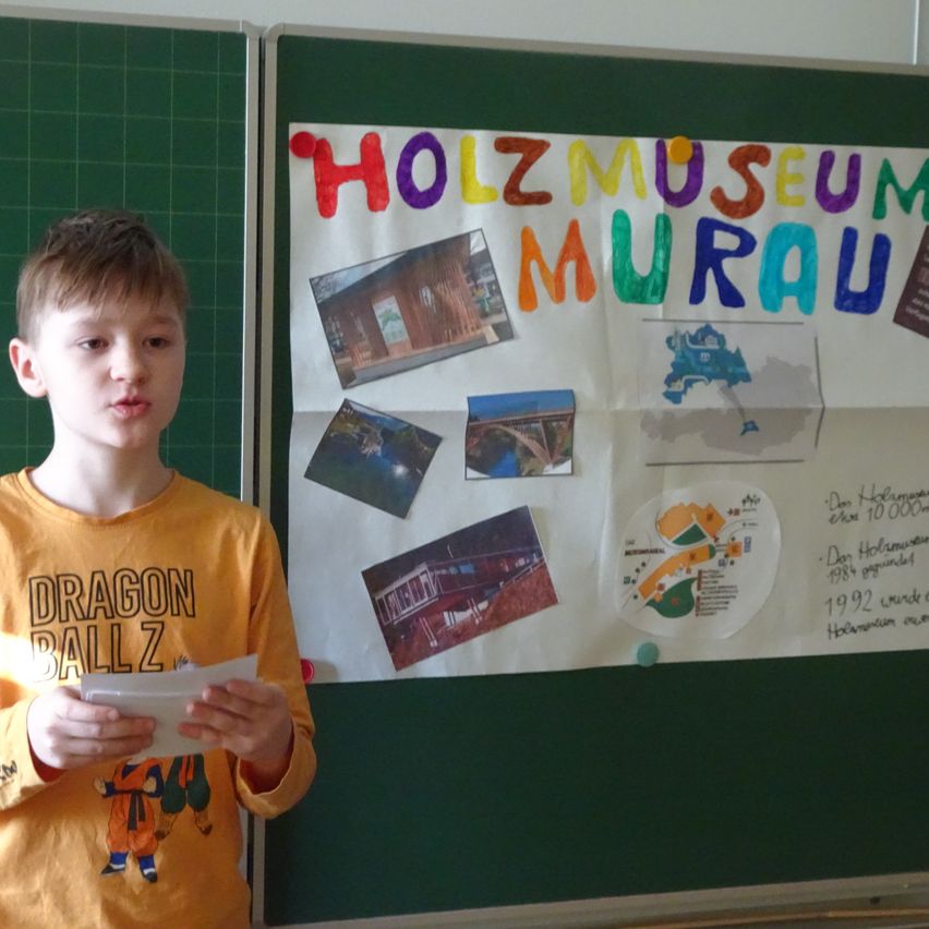 Ein Junge in einem gelben T-Shirt mit einem Comic-Charakter hält ein Blatt Papier vor einem Bulletin Board mit einem farbenfrohen Poster mit dem Titel Holzmuseum.