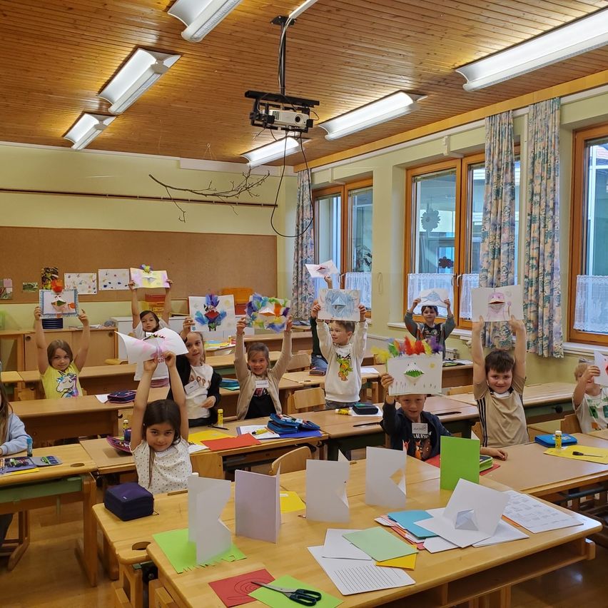 Kinder in einem Klassenzimmer halten ihre fertigen Kunstprojekte hoch und lächeln. Die Räume sind hell, und das Sonnenlicht dringt durch die Fenster.