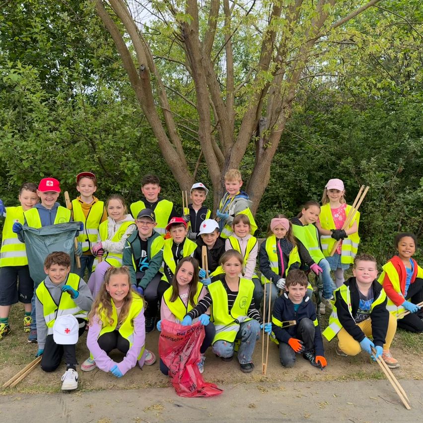 Eine Gruppe von Kindern in gelben Westen und Handschuhen posiert für ein Foto im Freien, hält Stöcke und Müllsäcke in der Hand, mit einem Baum und Grün im Hintergrund.