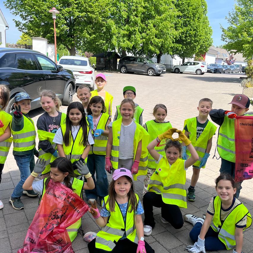 Eine Gruppe junger Kinder in gelben Westen steht und hockt draußen auf einem Parkplatz, mit Bäumen und geparkten Autos im Hintergrund.