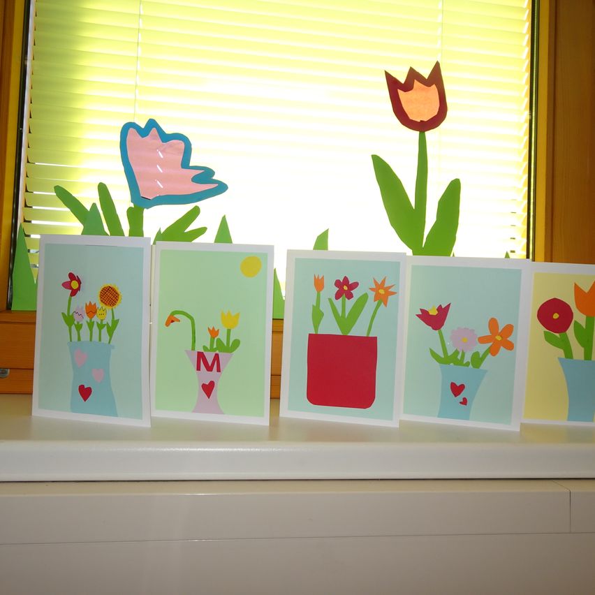 Fünf handgefertigte Karten mit farbenfrohen Blumen in Vasen sind auf einem Fenstersims ausgestellt. Die Karten sind mit verschiedenen Blumen wie Tulpen und Gänseblümchen verziert und vor einem Hintergrund mit weißen Fenstern und gelben Jalousien platziert.