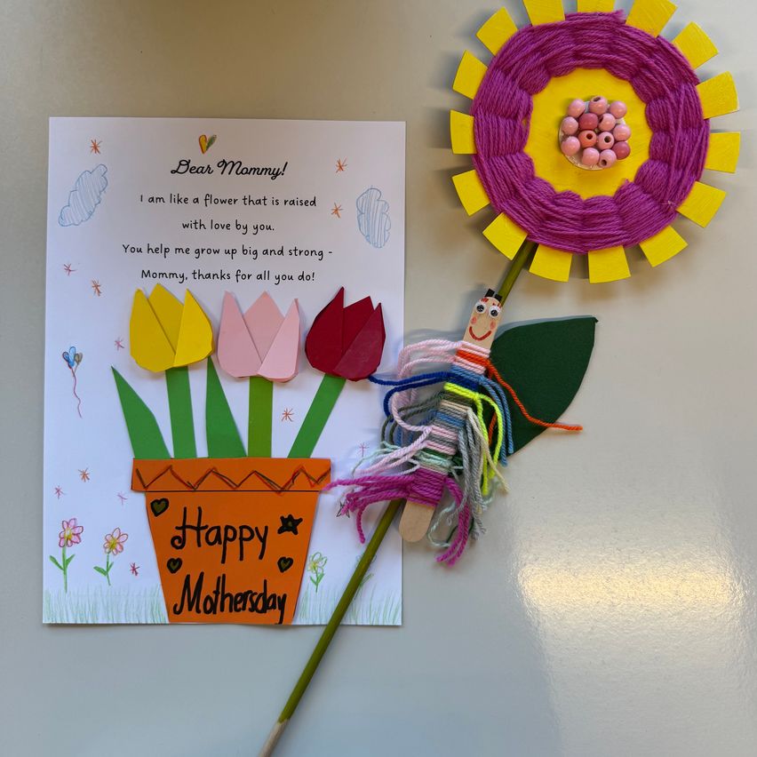 Eine handgefertigte Karte mit Blumen und einer Nachricht, 'Liebe Mama! Ich bin wie eine Blume, die mit Liebe von dir aufgezogen wurde.' Die Karte ist an einem farbenfrohen Stock befestigt, der ein Blumenende hat.