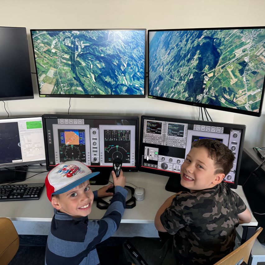 Two boys sit at a flight simulator station with multiple monitors and control panels. One holds a control stick, both smiling.