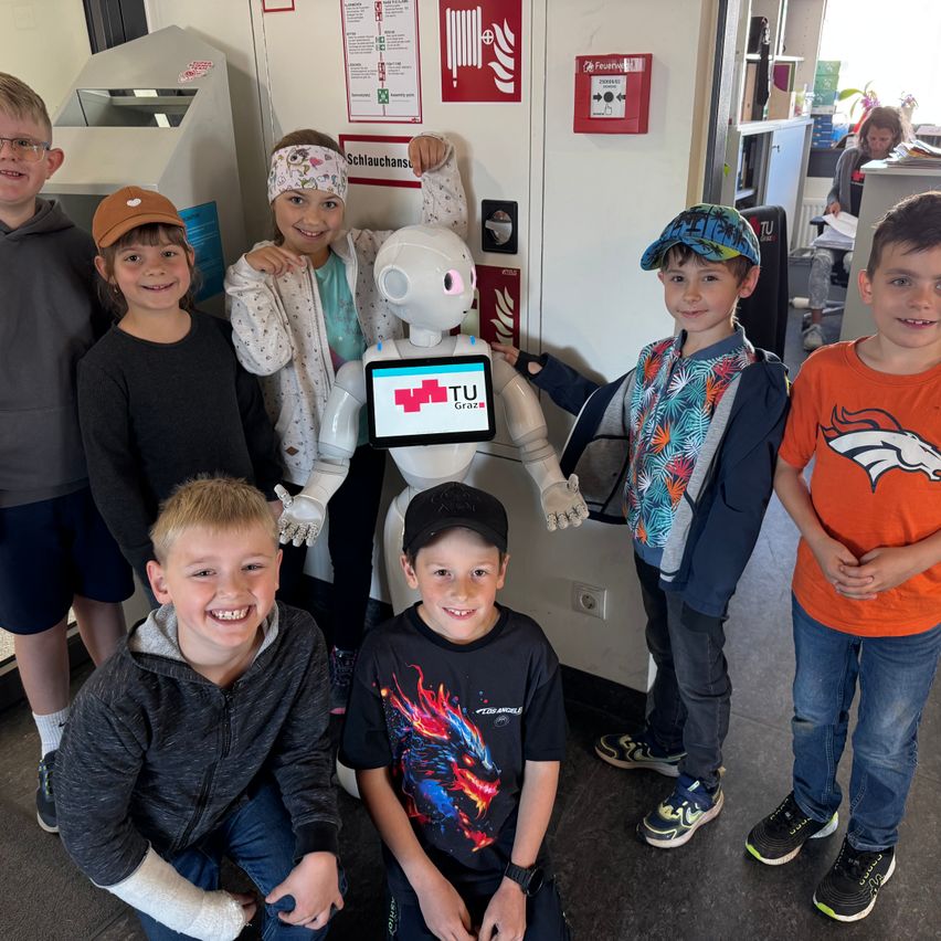 Kinder posieren mit einem weißen Roboter. Der Roboter hat einen Bildschirm mit TU Graz Logo. Sie stehen in einem Raum mit Feuerlöschern und Sicherheitszeichen.