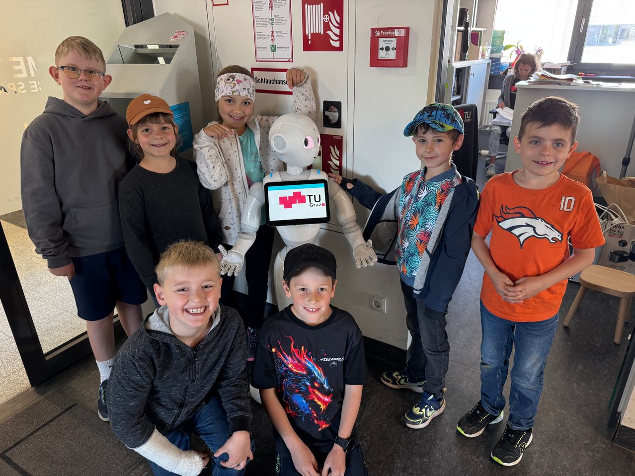 Children posing with a white robot. The robot has a screen with TU Graz logo. They are standing in a room with fire extinguishers and safety signs.