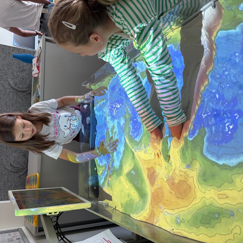 Zwei junge Mädchen spielen mit einer großen farbenfrohen interaktiven Anzeige in einem Klassenzimmer. Eine lehnt sich über die Anzeige, während die andere mit ihr interagiert.