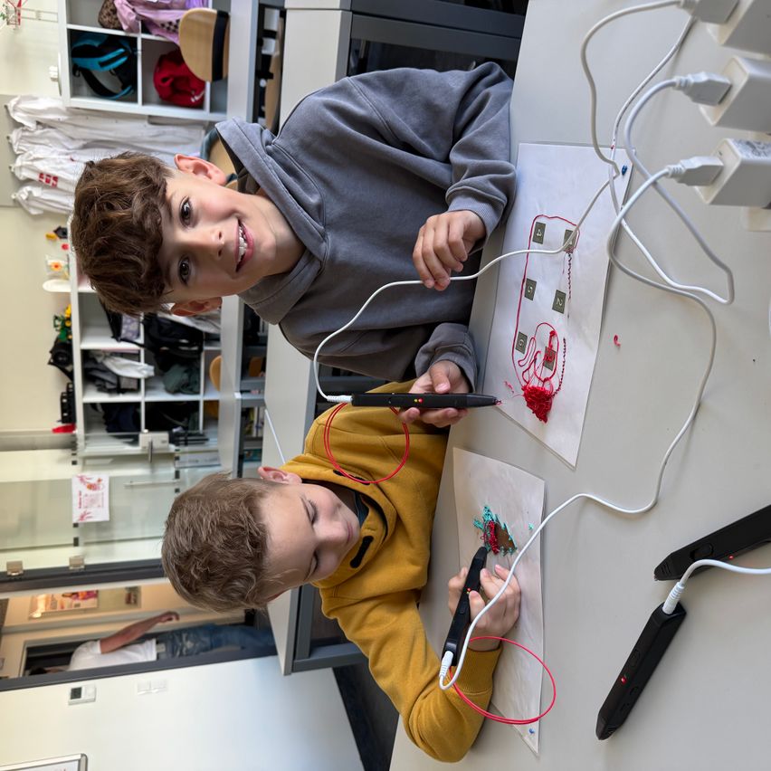 Two young boys work on a project with wires and electronic equipment on a white desk. One boy smiles while looking at a diagram on paper.