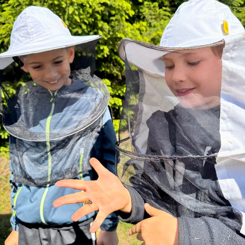 Zwei Kinder in Imkeranzügen und Hüten sind draußen. Eines hält eine Biene. Sie stehen vor grünen Büschen.