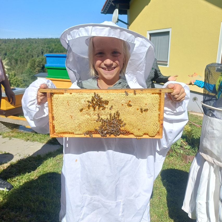 Ein junges Mädchen in einem Imkeranzug lächelt und hält ein bienenvolles Honigwaben-Fach draußen. Dahinter befinden sich Bienenstöcke, ein Gebäude und Bäume.