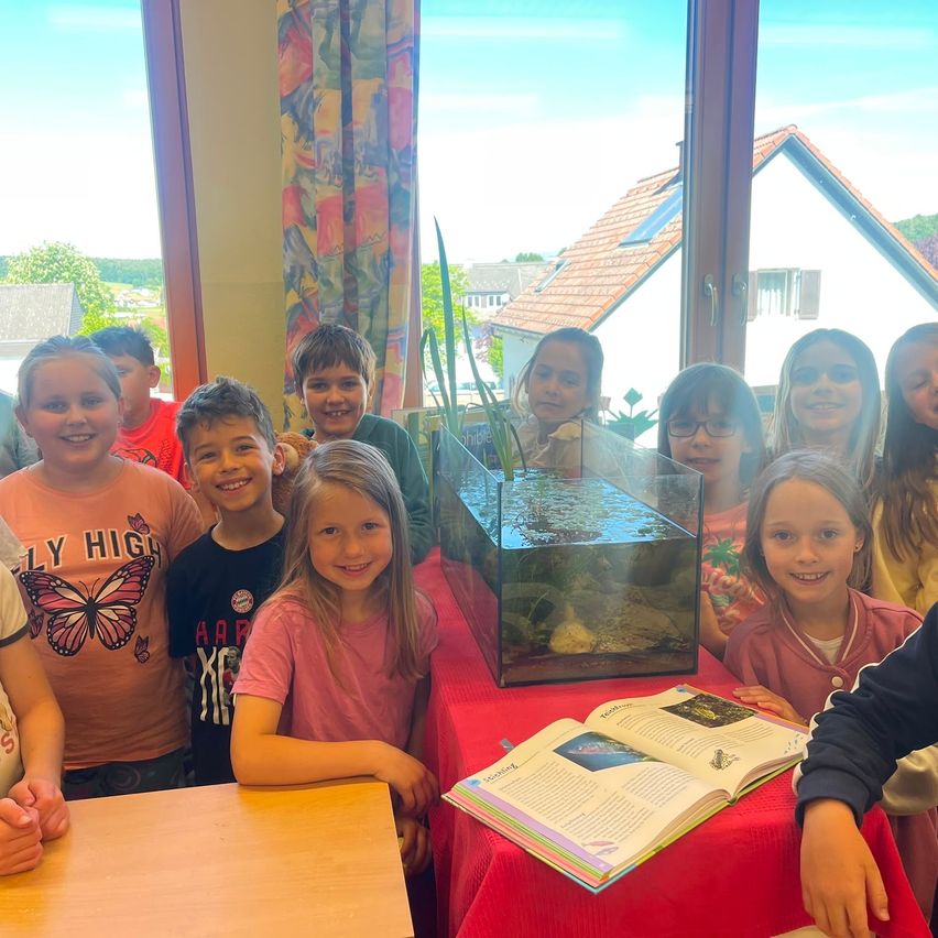 Eine Gruppe von Kindern ist um ein Aquarium mit einer Pflanze darin versammelt und lächelt für ein Foto. Ein Buch liegt offen auf einem roten Tisch vor ihnen.