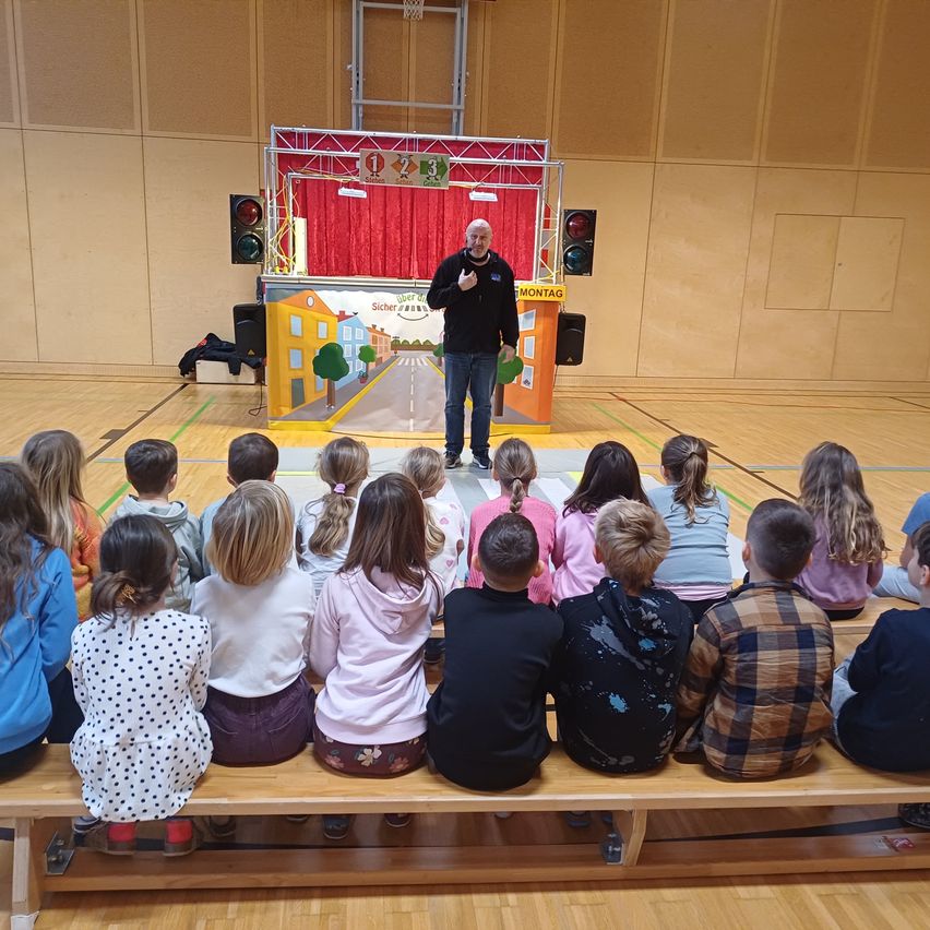 A man stands on a stage with a traffic light setup, addressing a group of children sitting on a bench in front of him in a gymnasium.