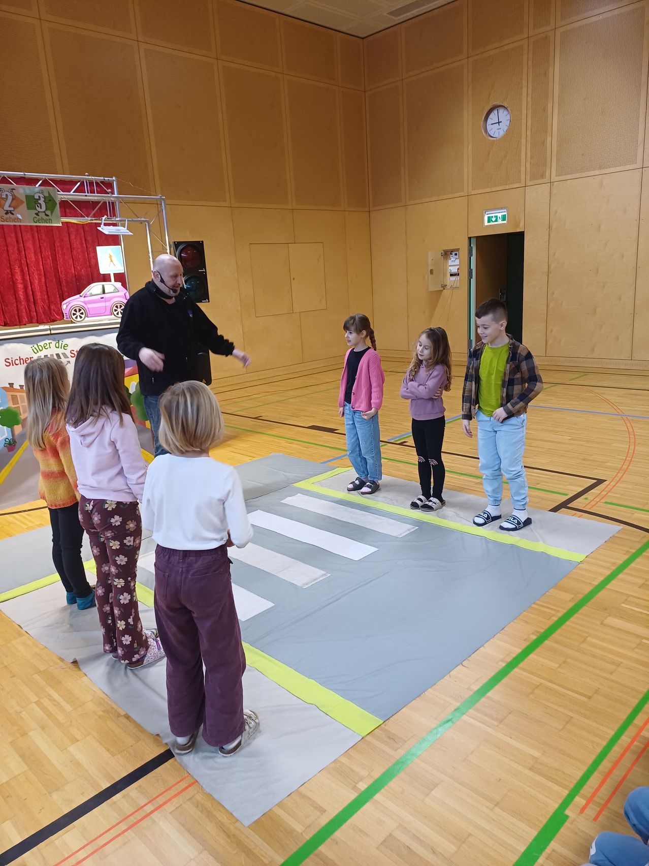 A group of children and a man are standing in a gym. The children are standing on a large gray mat that resembles a crosswalk. The man is speaking to the children.