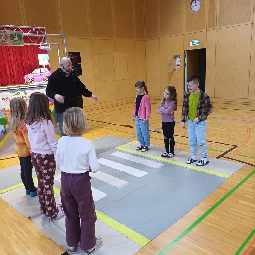 Eine Gruppe von Kindern und einem Mann stehen in einer Turnhalle. Die Kinder stehen auf einem großen grauen Teppich, der einem Zebrastreifen ähnelt. Der Mann spricht zu den Kindern.